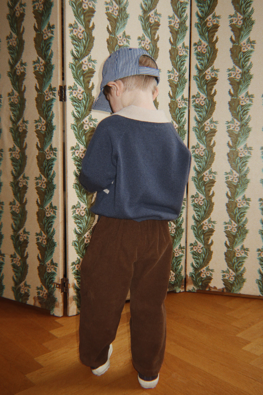 Child wearing a navy collared sweater, brown trousers and a striped cap